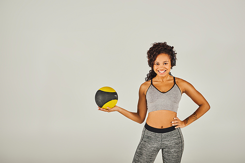 Curly African American sportswoman in active wear confidently holds a yellow and black ball in a studio setting.