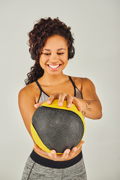 Curly African American sportswoman in active wear energetically holding a yellow and black exercise ball in a studio.