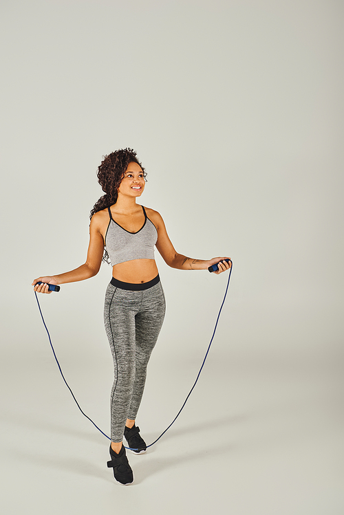 Curly African American sportswoman in active wear jumps with a skipping rope on a white background.