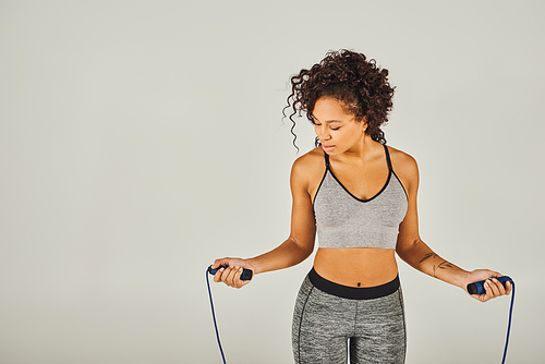 A curly African American sportswoman in active wear holding a skipping rope in front of a gray background.