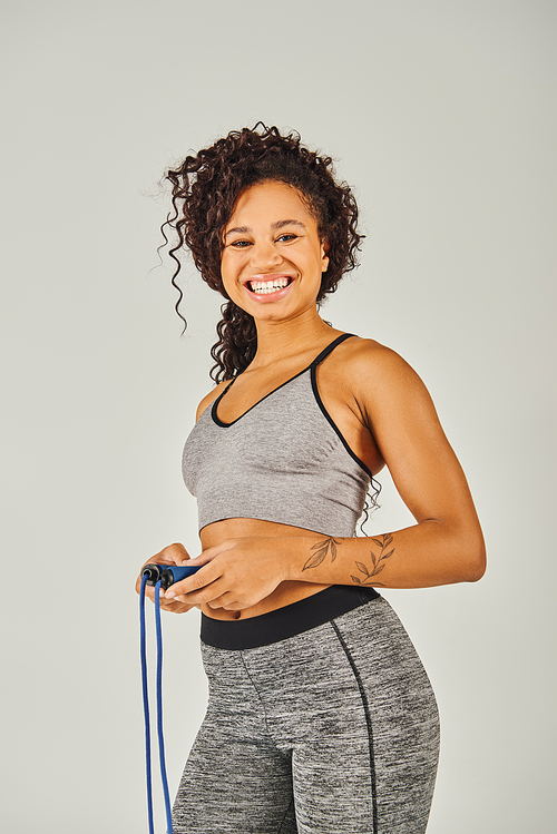 A curly African American sportswoman in activewear smiles while holding a skipping rope in a studio against a grey background.