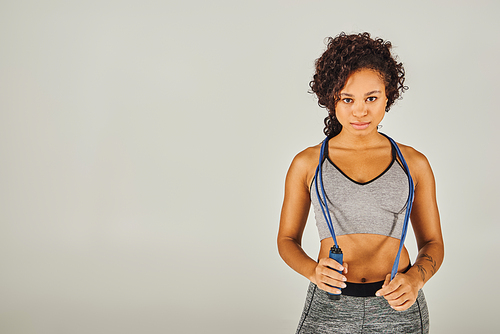 Curly African American sportswoman in active wear exuberantly holding a jump rope in a studio with a grey background.