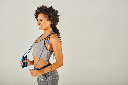 Curly African American woman in activewear holding a jump rope in a studio with grey background.