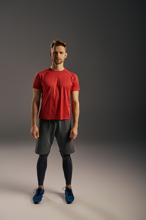 A man in a red t-shirt and grey shorts confidently strikes a pose in front of a serene grey background.