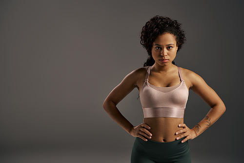 Curly African American sportswoman in a sports bra, confidently posing in a studio with a grey background.