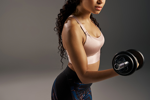 A curly-haired African American sportswoman in activewear works out with dumbbells in a grey studio background.