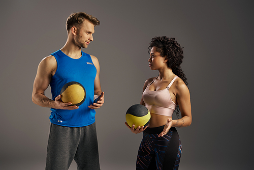 Young multicultural and fit couple in active wear standing next to each other, holding exercise balls in a studio setting.