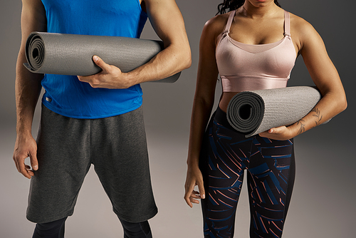 A young multicultural and fit couple in active wear holding a yoga mat in a studio setting against a grey background.