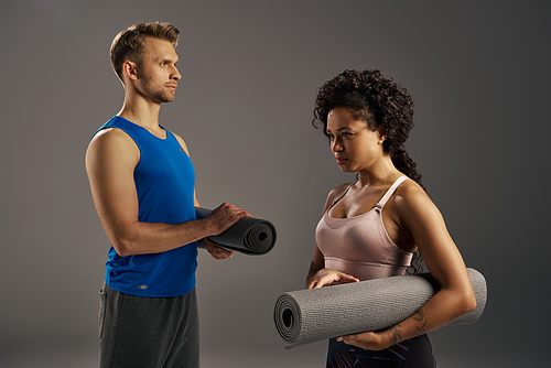 Multicultural couple in activewear stand next to a yoga mat, ready for a workout session in a studio on a grey background.