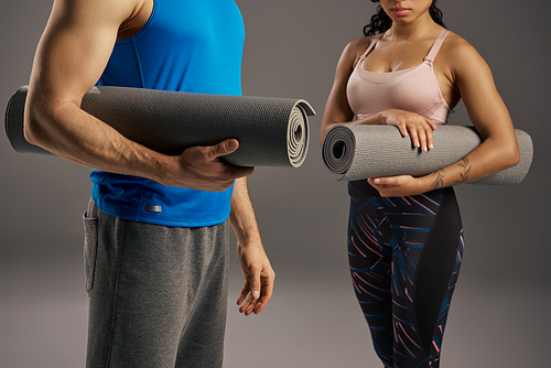 Young multicultural couple in active wear, holding a yoga mat in a studio setting against a grey background.