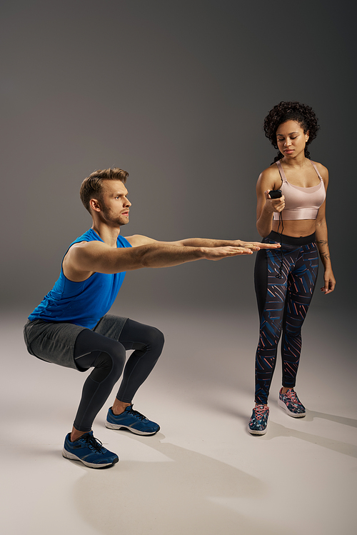 A young, multicultural couple in activewear perform squats on a grey background in a studio setting.