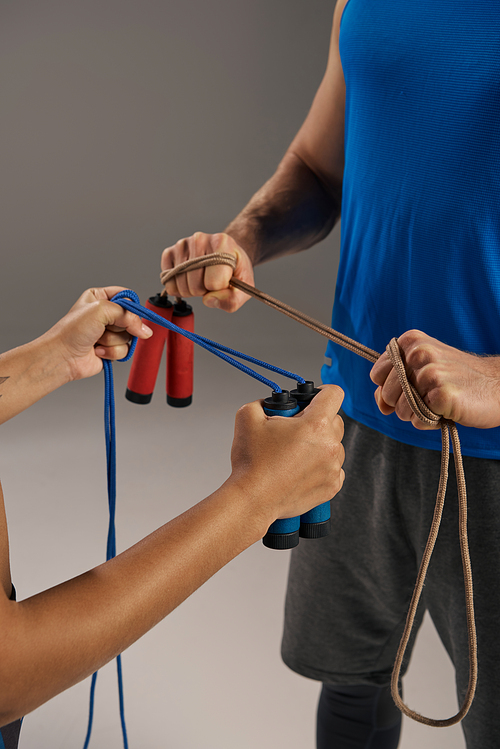 A man and a woman, both young and multicultural, in active wear, hold jump ropes in a fitness studio on a grey background.