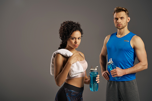 A multicultural, fit couple in active wear posing stylishly with water bottles on a grey studio background.