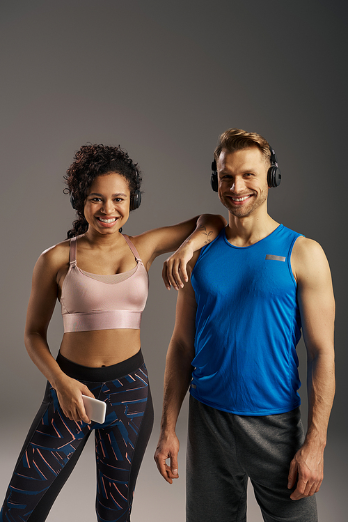 A fit multicultural couple in activewear strikes a pose together in a studio against a grey background.