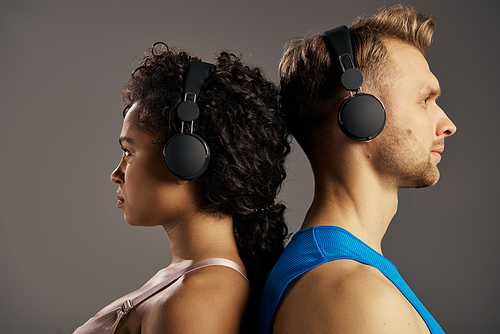 Young, multicultural couple in activewear, immersed in music, wearing headphones while working out in a studio against a grey backdrop.