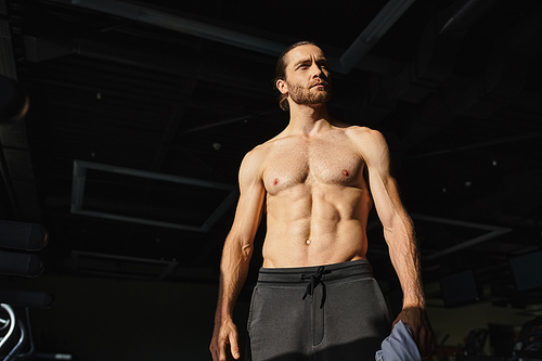 A muscular man in a dark space, shirtless and standing confidently.