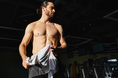 Muscular man in gym standing shirtless and holding towel in dark gym