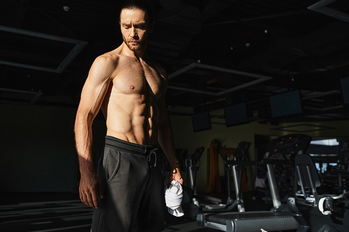 A muscular man, shirtless, standing in a gym setting, showcasing his physical strength and dedication to working out.