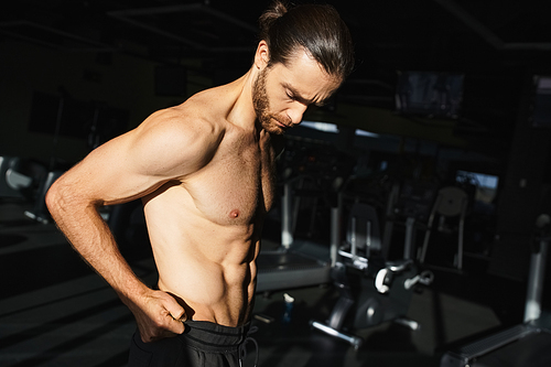 A shirtless man stands confidently in front of a gym machine, showcasing his muscular physique.