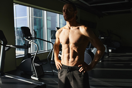 A muscular man without a shirt standing in front of a gym machine, focused and ready to workout.