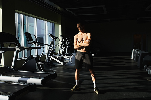 A muscular man without a shirt standing confidently in front of a row of treadmills in a gym.