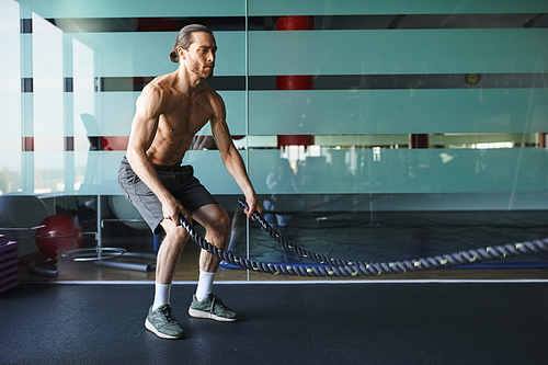 A muscular man without a shirt is intensely holding a battle rope in a gym, showcasing his strength and determination.