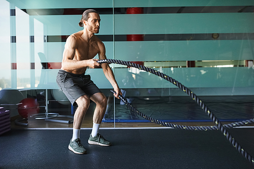 A shirtless, muscular man confidently holds a battle rope in a gym, showcasing his strength and determination.