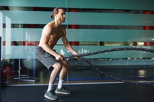 A muscular man without a shirt is gripping and pulling a battle rope in a gym, showcasing his strength and determination.