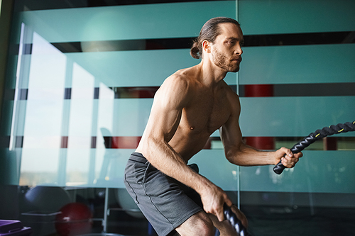 A shirtless, muscular man confidently holds a battle rope in a gym setting.