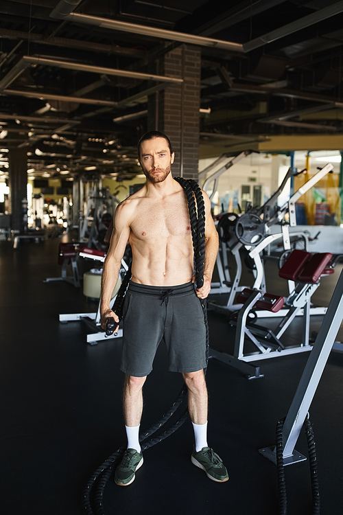 A muscular man without a shirt, standing in a gym, showcasing his dedication to fitness and strength training.