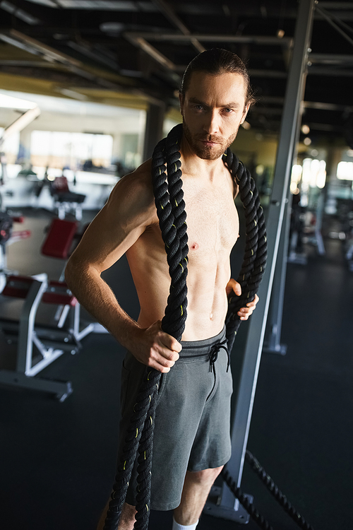 Shirtless man showcasing muscular physique while gripping a rope in gym.