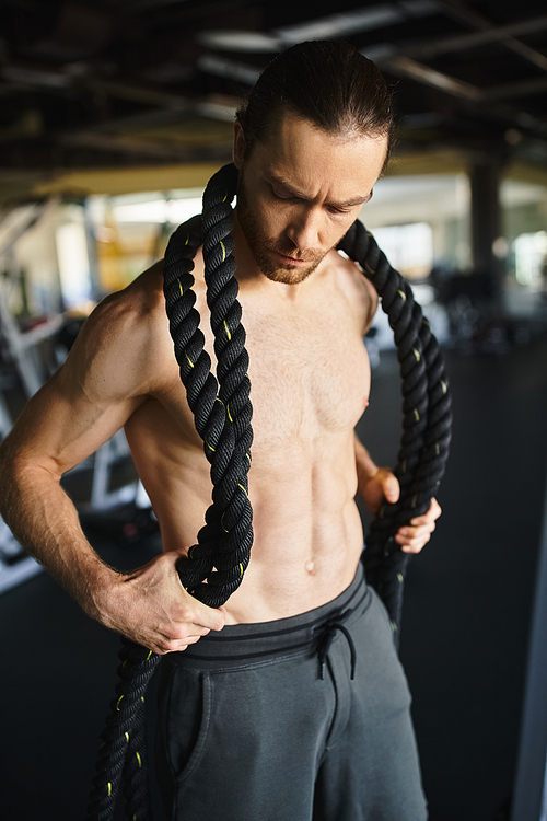 A shirtless muscular man holding a rope around his neck and body while working out in a gym.