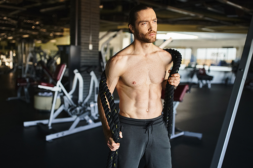 A shirtless, muscular man intensely focused on his gym workout with heavy ropes.