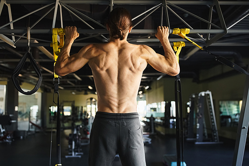 A shirtless man in a gym performs pull ups, showcasing his muscular frame and dedication to fitness.