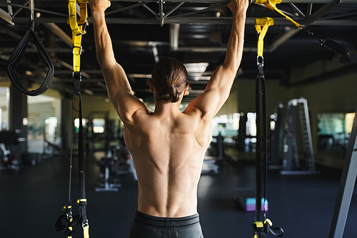 A shirtless muscular man is executing pull ups with technique and strength in a gym filled with exercise equipment.