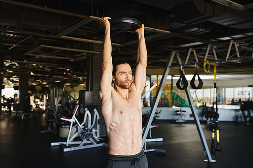 Muscular man without a shirt doing a pull up in a gym setting.