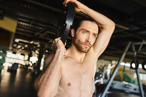 A shirtless man grips a weight plate in a gym, showcasing his muscular physique as he focuses on his workout routine.