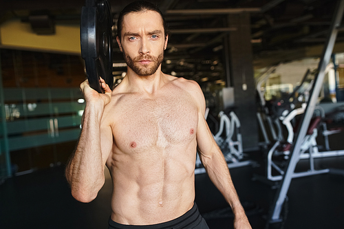 Shirtless man showcasing strength, holding a black weight plate in gym.