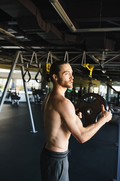 Shirtless man showcasing his strength while holding a weight plate in a gym setting.