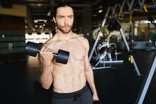 A shirtless man displaying his immense strength, holding two dumbbells, in the intense atmosphere of a gym.