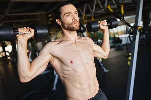 A muscular man without a shirt holding two dumbbells in a gym, showcasing his workout routine and physical strength.