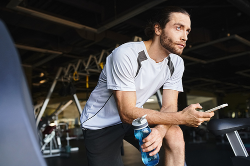 Muscular man sitting on a bench, holding a bottle of water and smartphone