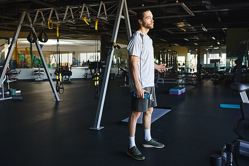 A muscular man holding a bottle of water, stands confidently in a gym.