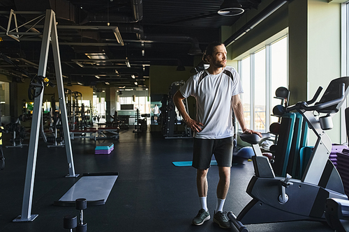 A muscular man standing in a gym surrounded by a rack of exercise equipment.