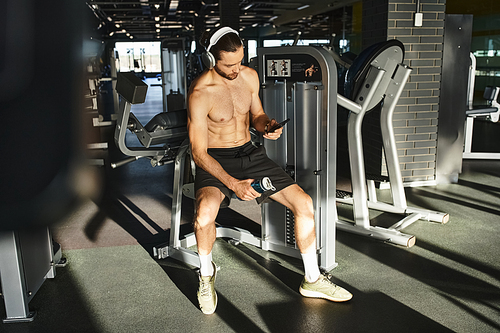 A shirtless man sitting on a bench, engrossed in his cell phone and headphones