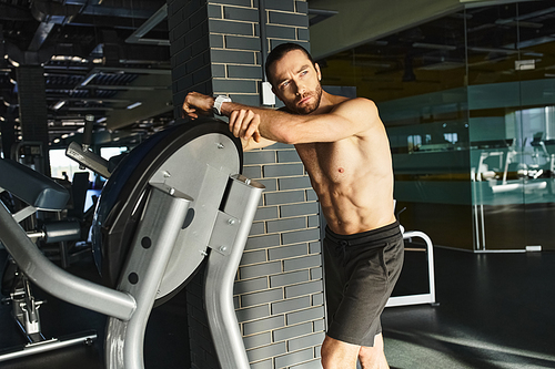 A shirtless, muscular man leans against a wall next to a workout machine in a gym setting.