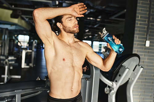 Strong shirtless man taking a break, holding a bottle of water in a gym setting.