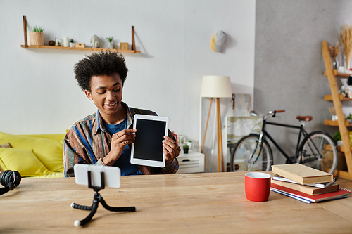 A young African American male blogger is sitting at a table with a tablet in front of him, discussing into a phone camera.