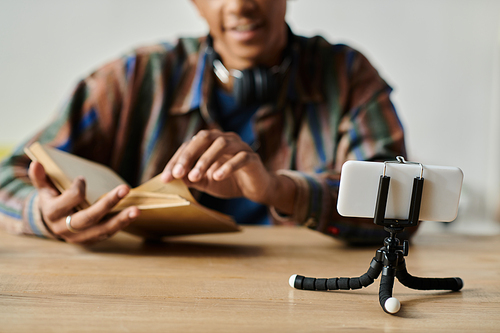 African American man reads book while vlogging on phone tripod.