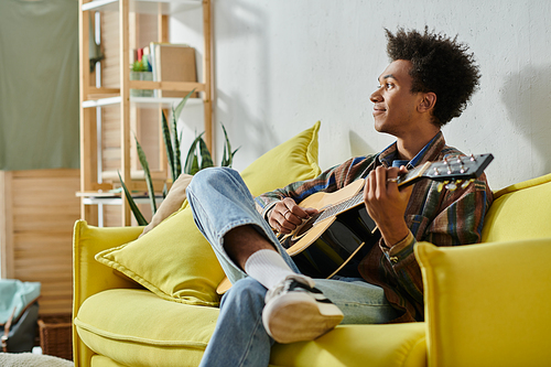 A young African American man plays an acoustic guitar while seated on a yellow couch.